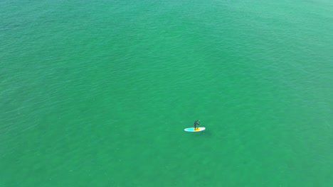 Paddle-Boarder-Isolated-on-Turquoise-Waters-at-Constantine-Bay-in-Cornwall-with-an-Aerial-Drone-Orbiting,-UK