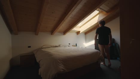 Mann-Kommt-Ins-Schlafzimmer,-Betrachtet-Das-Sonnenlicht-Aus-Dem-Velux-Dachfenster-Und-Setzt-Sich-Dann-Auf-Das-Bett