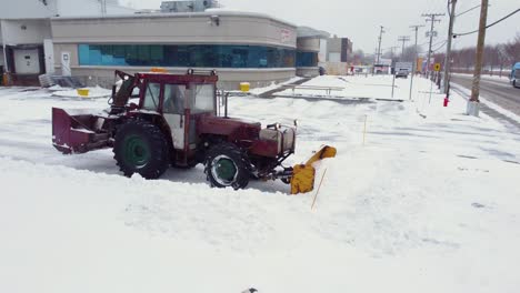 Roter-Traktor-Räumt-Schnee-Mit-Einem-Schneepflug-Auf-Einem-Büroparkplatz,-Mit-Schwenkaufnahme-Einer-Drohne-Aus-Der-Luft
