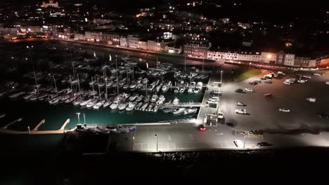 Short-high-drone-flight-at-night-over-QE-II-Marina,-St-Peter-Port,-Guernsey-with-slipway,-Glategny-Esplanade-and-boats-in-their-moorings-and-twinkling-lights-of-town-as-a-backdrop