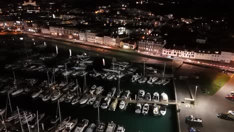 Drone-flight-at-night-over-QE-II-Marina,-St-Peter-Port,-Guernsey-with-slipway,-Glategny-Esplanade-and-boats-in-their-moorings-and-twinkling-lights-of-town-as-a-backdrop