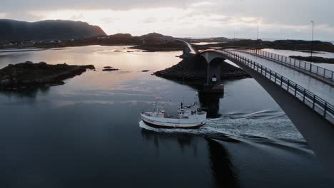 Aerial-view,-pushing-in,-tilting-down,-towards-a-fisher-boat-returning-to-port-under-the-Flakstadbruene-bridge,-near-Fredvang,-Lofoten-Islands,-Norway