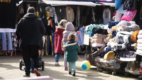 Shoppers,-including-children-with-colorful-balloons,-browse-through-an-outdoor-market-filled-with-clothing-and-blankets