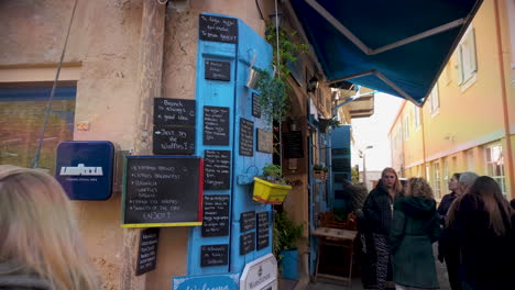 Eine-Lebhafte-Café-Szene-In-Lefkara,-Zypern,-Mit-Farbenfroher-Beschilderung,-Einer-Blauen-Tür-Und-Einer-Gruppe-Von-Menschen,-Die-Sich-Draußen-Versammelt-Haben