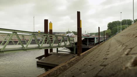 Statische-Aufnahme-Einer-Grünen-Brücke-über-Wasser-Mit-Bewölktem-Himmel-Und-Industrieller-Kulisse,-Hölzerne-Vordergrunddetails