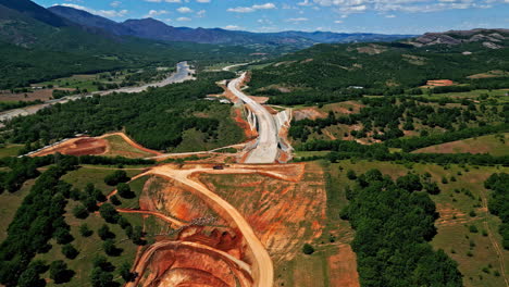 Highway-road-construction-site-through-magical-hilly-landscape,-aerial-drone-view