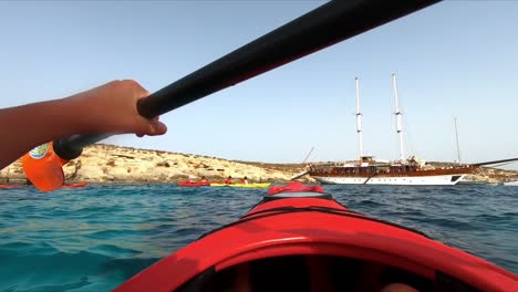 Paddling-in-the-Mediterranean-Sea-on-a-sunny-day-near-Comino's-shores,-heading-towards-a-moored,-grand-classic-sailing-ship-in-Malta