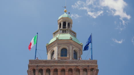 Italian-flags-waving-atop-Bologna's-historic-tower-under-blue-sky