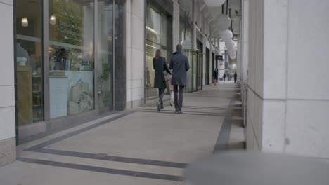 Two-people-walking-through-a-stylish-urban-arcade-lined-with-shops-and-restaurants,-captured-during-the-day