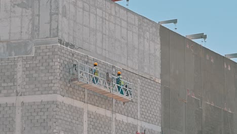 Workers-labouring-on-a-suspended-platform-at-a-construction-site-during-a-hot-summer-day-in-Dubai,-United-Arab-Emirates