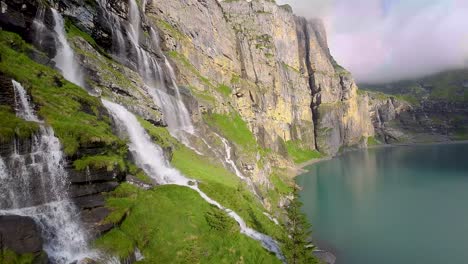 Luftflug-Neben-Einem-Wunderschönen-Großen-Wasserfall-In-Einer-Berglandschaft,-Drohne-Fliegt-über-Einen-Blauen-See---Oeschinensee,-Schweiz