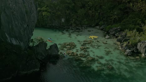 Tourists-kayak-in-still-water-by-Philippines-mountain-landscape,-aerial-push-in