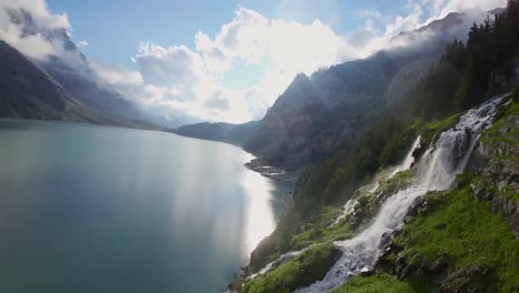 Aerial-flight-next-to-a-beautiful-big-waterfall-on-a-mountain-Landscape,-Drone-Flying-Over-a-Blue-Lake---Oeschinen-Lake,-Switzerland