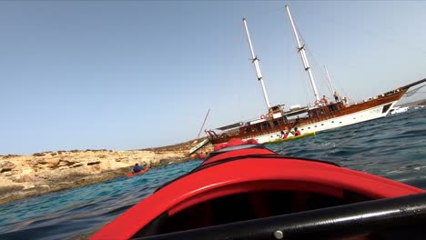 Un-Grupo-De-Personas-En-Kayak-Reman-Alrededor-De-Un-Velero-Tradicional-Maltés-En-El-Mar-Mediterráneo-Frente-A-La-Costa-De-Comino