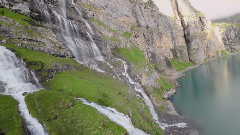 Luftflug-Neben-Einem-Wunderschönen-Großen-Wasserfall-In-Einer-Berglandschaft,-Drohne-Fliegt-über-Einen-Blauen-See---Oeschinensee,-Schweiz
