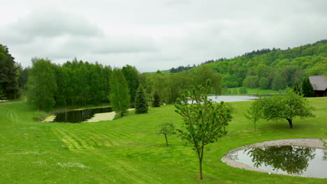 A-serene-aerial-view-of-a-manicured-park-with-small-ponds,-lush-greenery,-and-surrounding-forested-hills