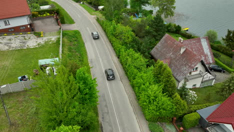 An-aerial-view-of-a-road-alongside-a-lake,-with-houses-and-green-foliage-lining-the-sides-with-cars-on-the-road
