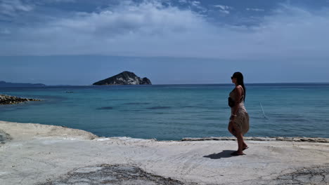 Woman-walking-on-a-serene-beach-with-a-distant-island-and-a-calm-blue-sea-under-a-partly-cloudy-sky