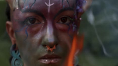 A-person-with-colorful-face-paint-designed-for-the-Scottish-Pagan-holiday-Beltane-and-torch-stares-intently-as-smoke-swirls-around-them