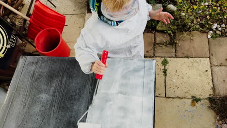 A-young-boy,-equipped-with-protective-goggles,-uses-a-paint-roller-to-paint-a-table-outdoors