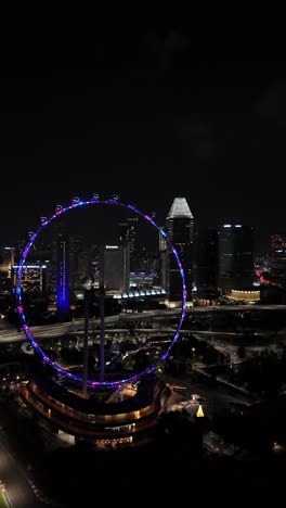 Antena-Nocturna-Giratoria-De-La-Noria-Bellamente-Iluminada-De-Singapur-En-El-área-De-Marina-Bay-Sands,-Vista-Aérea-Vertical