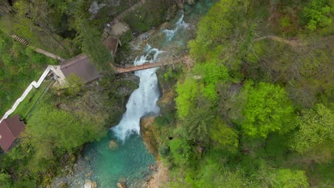 Aerial-view-of-a-vibrant,-turquoise-river-with-a-waterfall-flowing-through-lush-greenery-and-a-wooden-bridge-crossing-over-it
