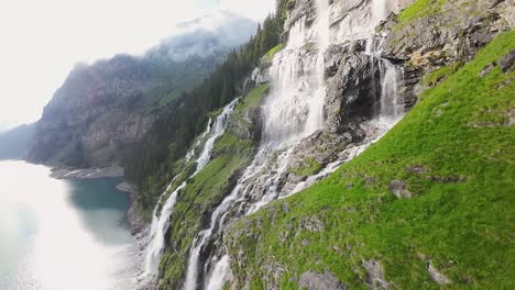Aerial-flight-next-to-a-beautiful-big-waterfall-on-a-mountain-Landscape,-Drone-Flying-Over-a-Blue-Lake---Oeschinen-Lake,-Switzerland