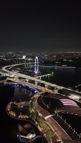 Impresionante-Antena-Nocturna-De-La-Zona-Iluminada-De-Marina-Bay-Sands-Con-La-Famosa-Noria-De-Singapur,-Iluminada-En-El-Cielo-Nocturno,-Vídeo-Aéreo-Vertical