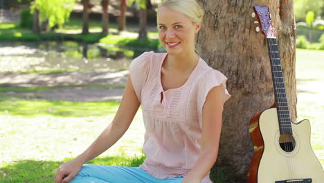 A-woman-sits-by-a-tree-with-her-guitar-as-she-smiles-at-the-camera