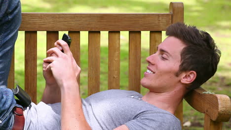 Man-lies-on-a-bench-while-looking-at-at-his-mobile-phone-before-laughing-and-looking-at-camera