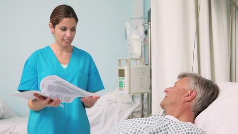 Lächelnde-Krankenschwester-Im-Gespräch-Mit-Einem-Patienten,-Der-Auf-Einem-Bett-Liegt-Und-Dabei-Ein-Papier-Hält