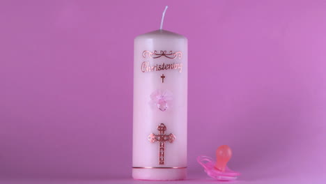Pink-pacifier-falling-beside-baptism-candle-on-pink-background