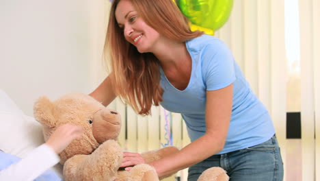 Woman-giving-a-teddy-bear-to-a-girl-in-a-bed-with-balloons
