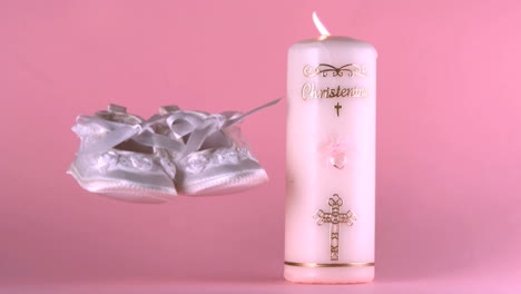 Baby-shoes-falling-beside-lit-baptism-candle-on-pink-background