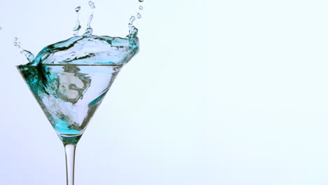 Ice-falling-into-cocktail-glass-of-blue-alcohol-on-white-background