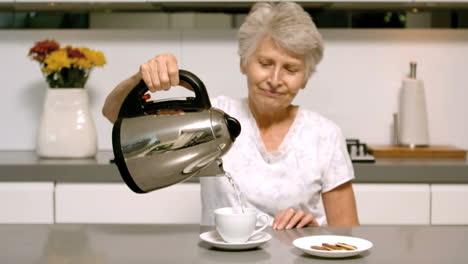 Elderly-woman-pouring-boiling-water-from-kettle-into-cup-in-kitchen