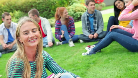 Estudiante-Rubia-Sonriendo-A-La-Cámara-Con-Amigos-Detrás-De-Ella-En-El-Césped