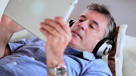 Hombre-Maduro-Tumbado-En-El-Sofá-Usando-Su-Tableta-Digital-Escuchando-Música