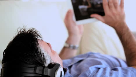 Mature-man-lying-on-the-couch-listening-to-music-and-using-tablet