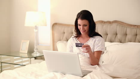 Schöne-Brünette-Mit-Ihrem-Laptop-Im-Bett-Sitzend,-Um-Online-Einzukaufen