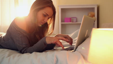 Pretty-happy-brunette-lying-on-bed-typing-on-laptop