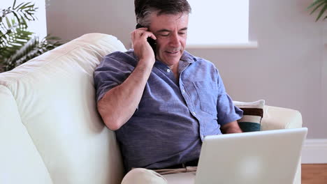 Mature-man-sitting-on-the-couch-using-laptop-and-answering-the-phone