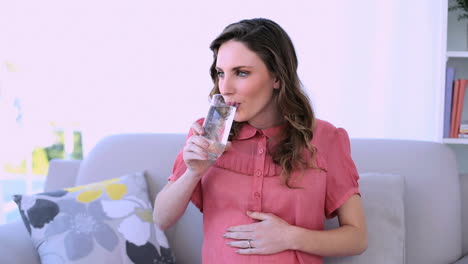 Modelo-Bastante-Embarazada-Sentada-En-Su-Sofá-Bebiendo-Un-Vaso-De-Agua