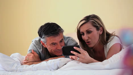 Smiling-couple-looking-at-smartphone-lying-on-bed