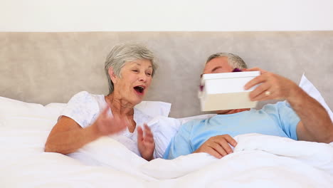 Happy-senior-couple-lying-in-bed-with-man-giving-a-gift