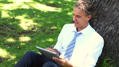 Happy-businessman-leaning-against-tree-using-tablet