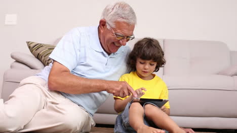 Senior-man-sitting-on-floor-with-his-grandson-using-tablet