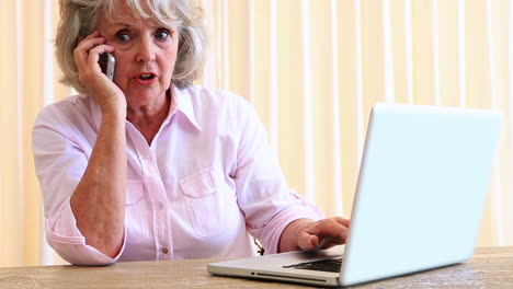 Ältere-Frau-Sitzt-Am-Tisch-Mit-Laptop-Und-Telefoniert