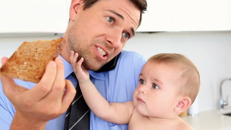 Businessman-holding-his-baby-and-talking-on-the-phone-while-eating-toast