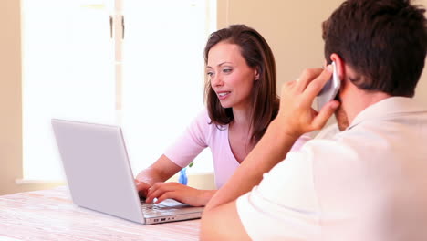 Woman-using-laptop-with-partner-talking-on-phone-at-the-table